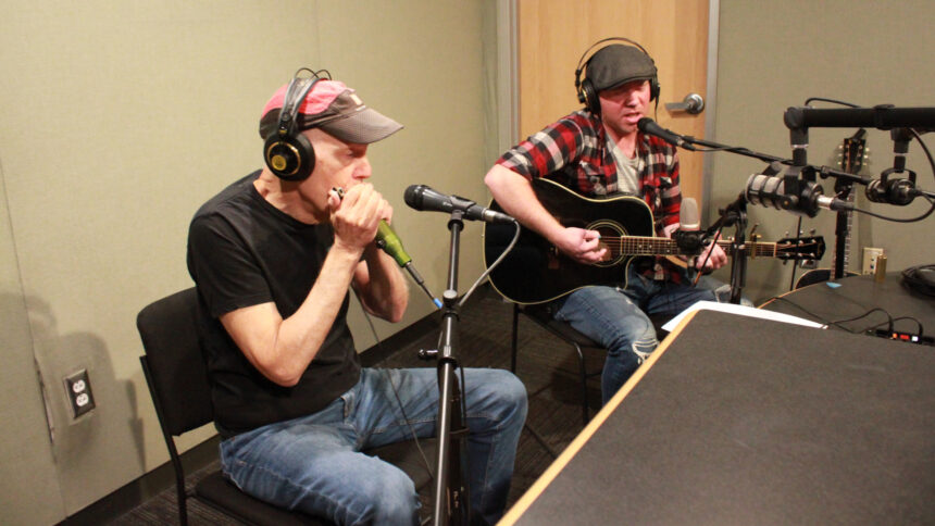 An adult wearing headphones playing harmonica into a microphone and an adult wearing headphones playing an acoustic guitar and singing into a microphone in a radio studio