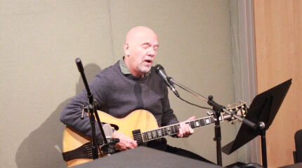 An adult playing an acoustic guitar and singing into a microphone in a radio studio