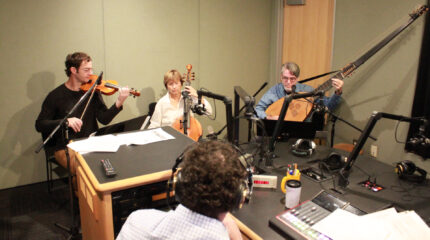 An adult playing a violin, an adult playing a viola de Gamba, and an adult playing a theorbo as an adult wearing headphones listens while sitting behind a microphone and a mixing board in a radio studio