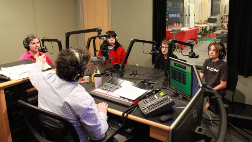 An adult wearing headphones, a teen wearing headphones, and two youth wearing headphones speaking into microphones being interviewed by an adult wearing headphones sitting behind a microphone and a mixing board in a radio studio