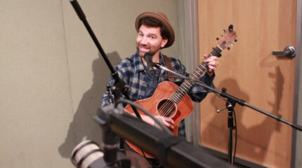 An adult smiling sitting behind a microphone and playing an acoustic guitar in a radio studio