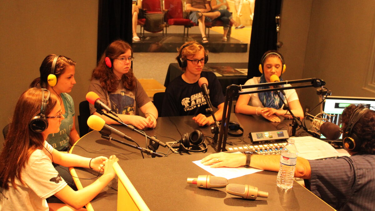 Five youth wearing headphones speaking into microphones being interviewed by an adult wearing headphones sitting behind a microphone in a radio studio