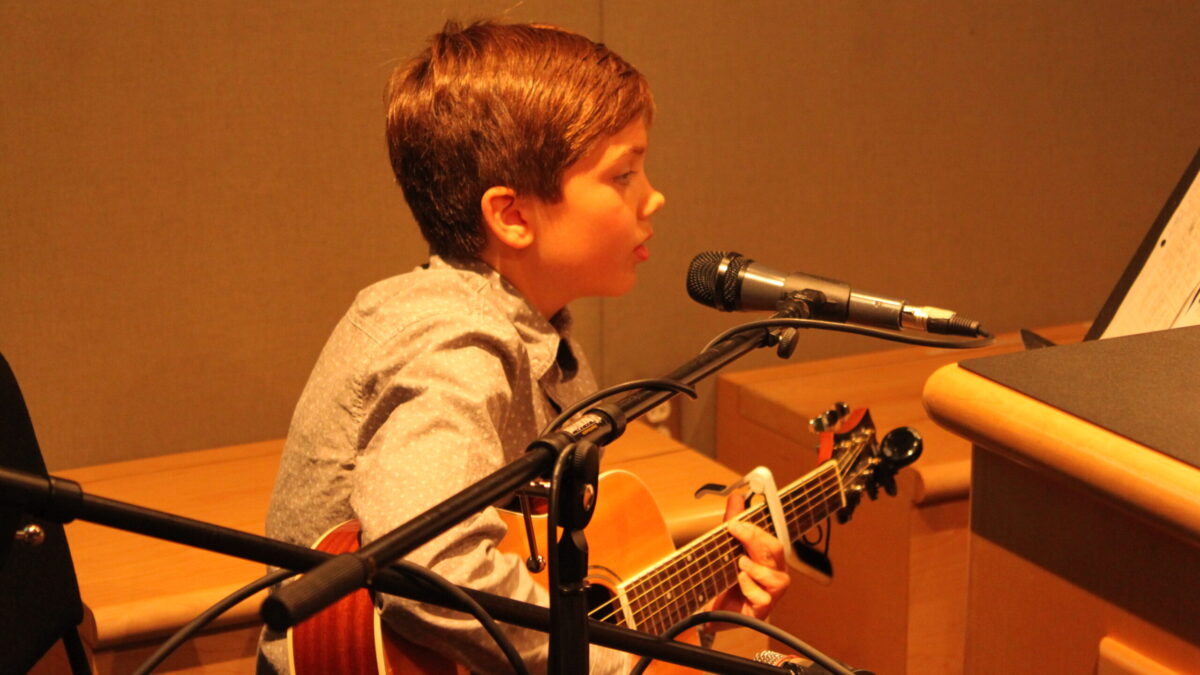 A youth singing into a microphone and playing an acoustic guitar in a radio studio