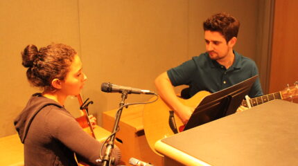 A youth and an adult playing acoustic guitars looking at a music stand in a radio studio