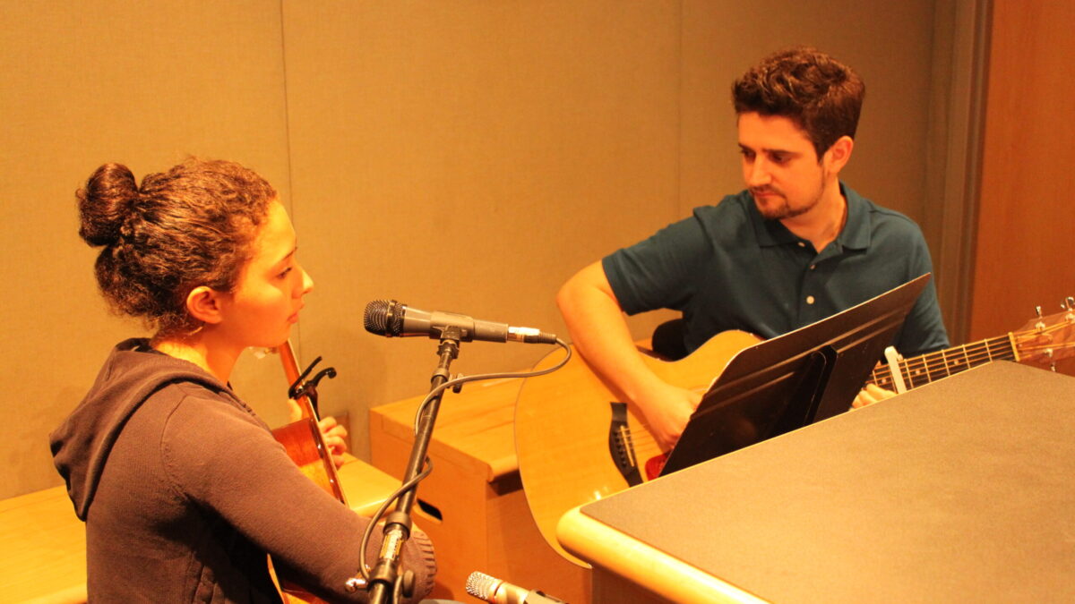 A youth and an adult playing acoustic guitars looking at a music stand in a radio studio