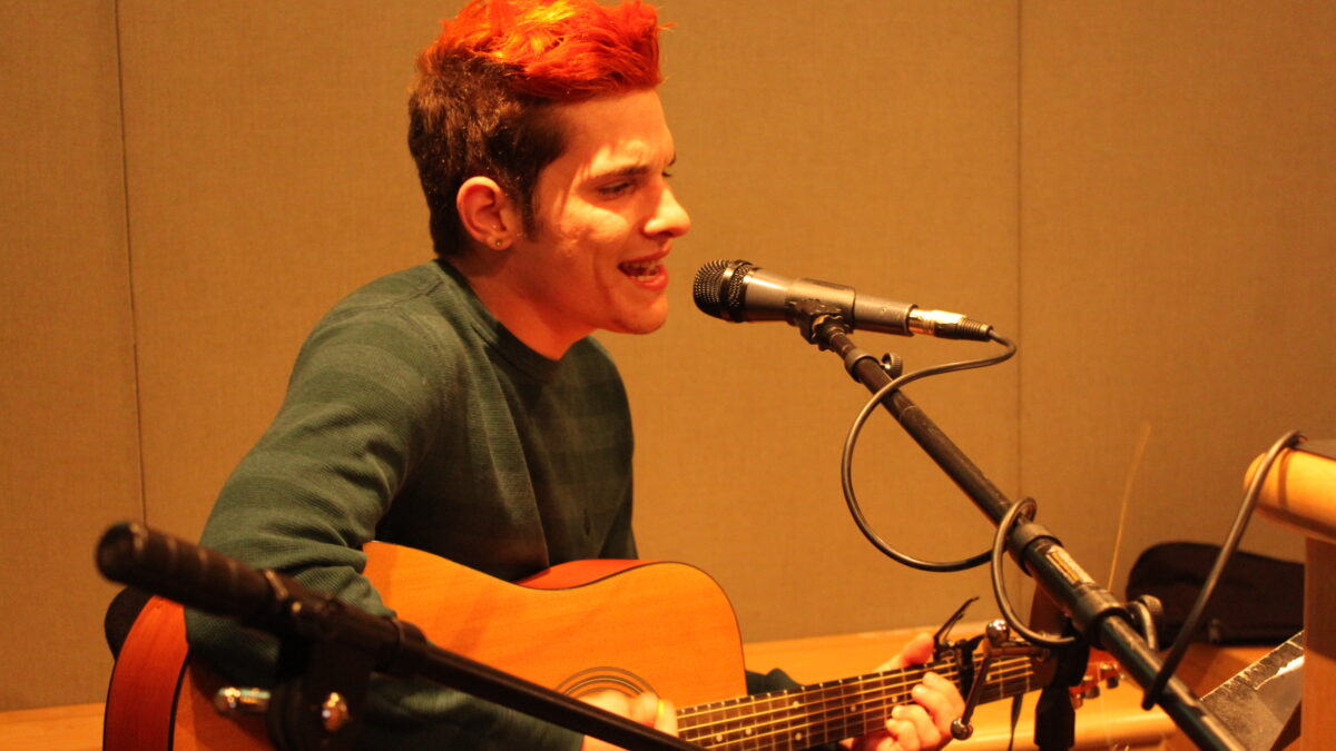 An adult singing into a microphone and playing an acoustic guitar in a radio studio