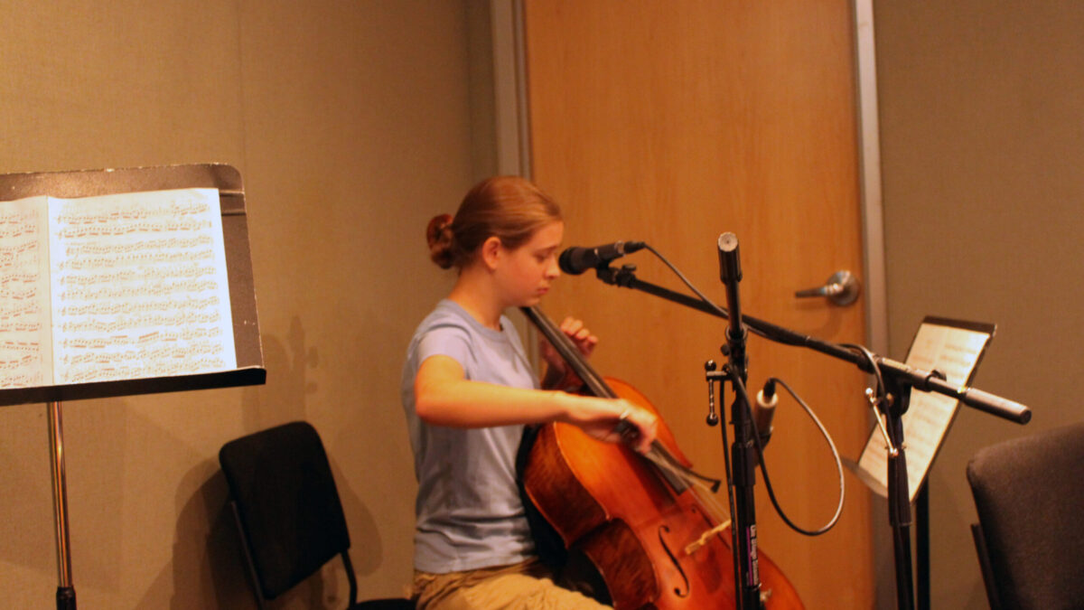 A youth playing a cello looking at sheet music on a music stand in a radio studio