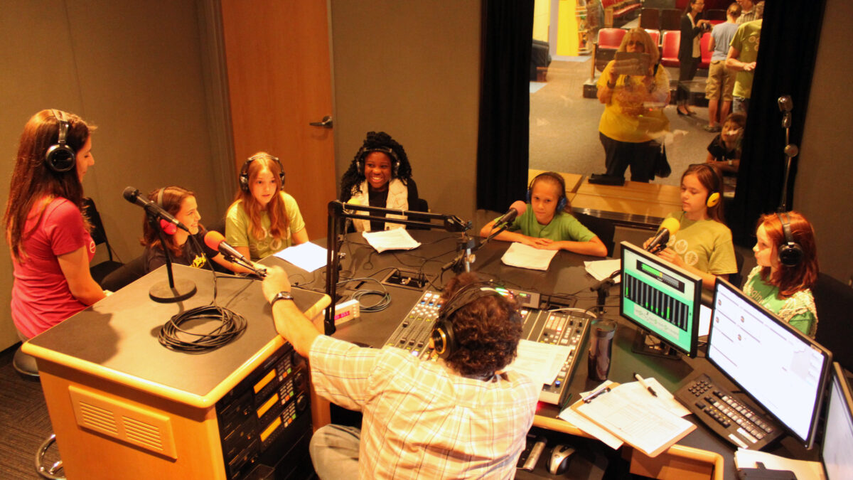 Six youth and an adult wearing headphones speaking into microphones being interviewed by an adult wearing headphones sitting behind a microphone and a mixer in a radio studio