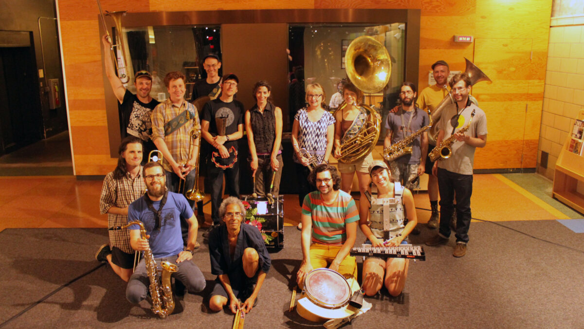 An adult kneeling with a trombone, an adult kneeling with a tenor saxophone, an adult kneeling with drumsticks, an adult kneeling with a snare drum, an adult kneeling with a glockenspiel, two adults standing posing with trombones, an adult posing with cymbals, an adult posing with a tambourine and a cowbell, an adult posing with a bass drum at their feet, an adult standing with a trumpet, an adult posing with a sousaphone, two adult standing with alto saxophones, and an adult posing with a tuba.