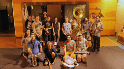 An adult kneeling with a trombone, an adult kneeling with a tenor saxophone, an adult kneeling with drumsticks, an adult kneeling with a snare drum, an adult kneeling with a glockenspiel, two adults standing posing with trombones, an adult posing with cymbals, an adult posing with a tambourine and a cowbell, an adult posing with a bass drum at their feet, an adult standing with a trumpet, an adult posing with a sousaphone, two adult standing with alto saxophones, and an adult posing with a tuba.