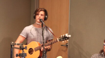 An adult wearing headphones singing into a microphone and playing an acoustic guitar in a radio studio