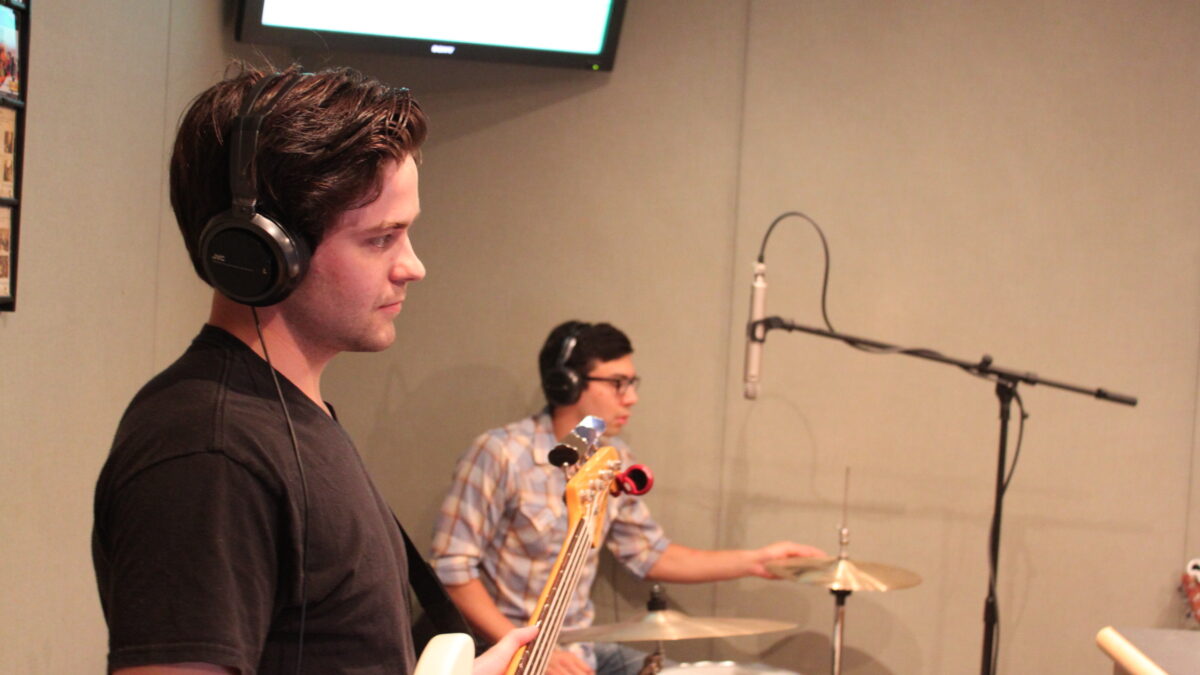 An adult wearing headphones holding an electric bass and an adult wearing headphones sitting at a drum set in a radio studio