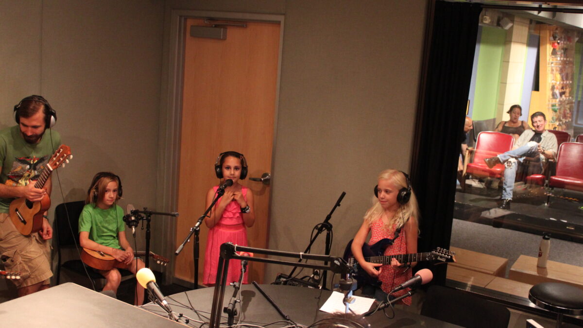IMG_9416 An adult holding a guitalele, a youth holding a guitalele, a youth standing behind a microphone, and a youth holding an electric guitar, all wearing headphones in a radio studio