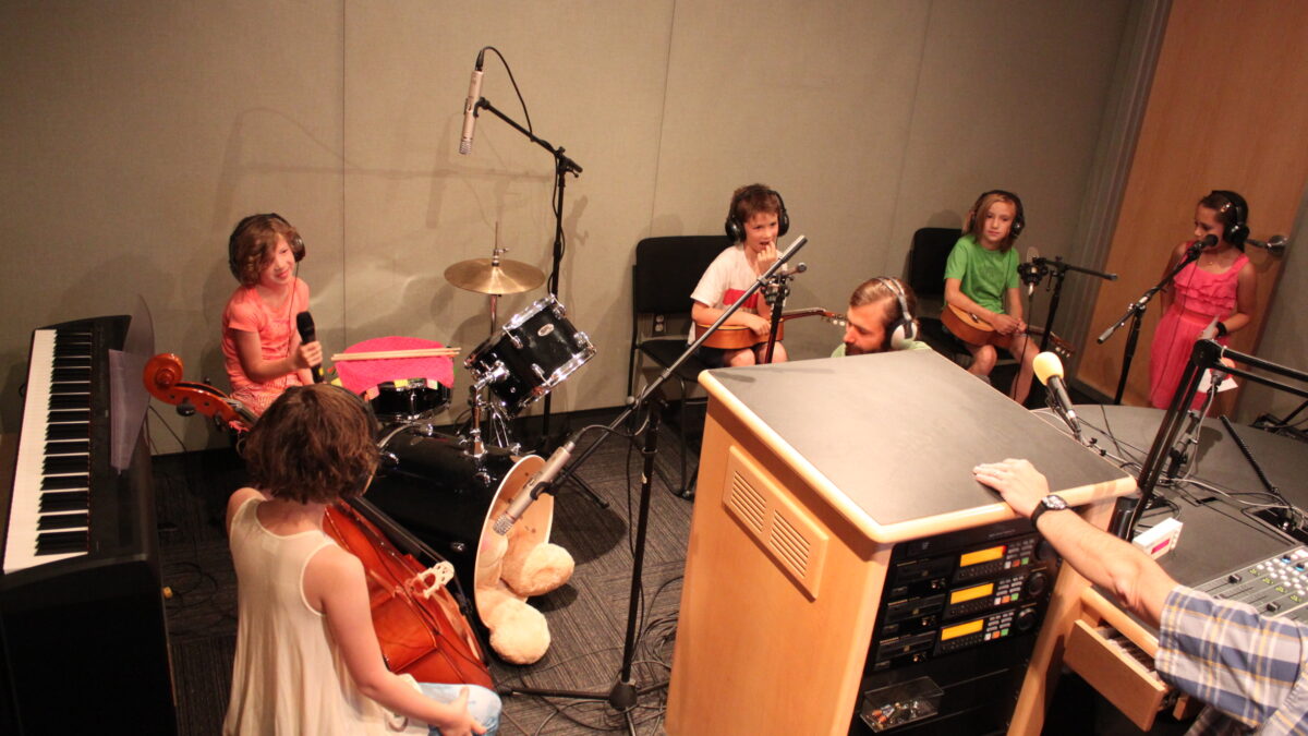 IMG_9417 A youth holding a cello, a youth sitting at a drum set holding a microphone, two youth holding guitaleles, and a youth standing behind a microphone, all wearing headphones in a radio studio