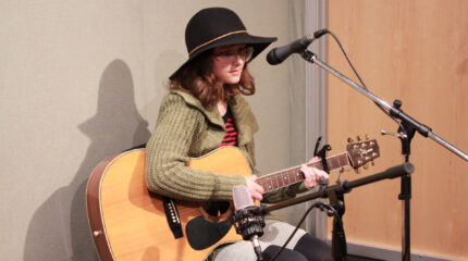 A teen playing an acoustic guitar singing into a microphone in a radio studio