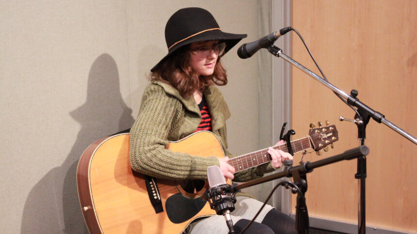A teen playing an acoustic guitar singing into a microphone in a radio studio