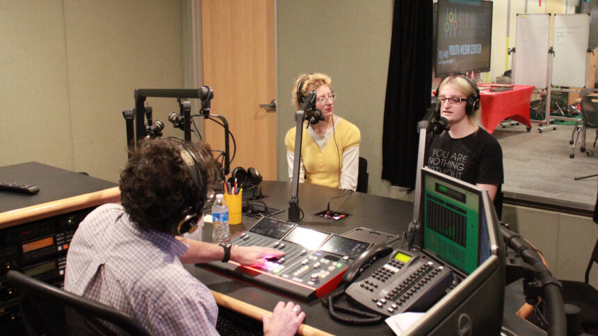 Two teens wearing headphones speaking into microphones being interviewed by an adult wearing headphones sitting behind a microphone and a mixing board in a radio studio