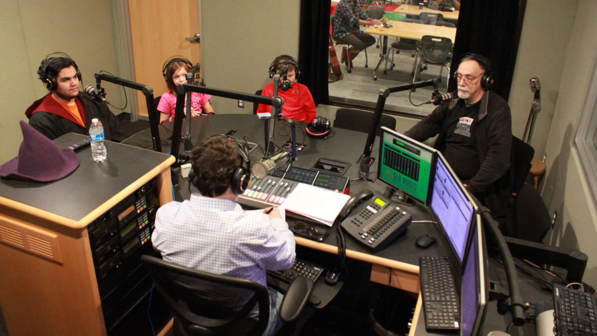 Society-of-American-Magicians-Youth-1 Three youth and an adult wearing headphones sitting behind microphones looking at an adult wearing headphones sitting behind a microphone and a mixing board writing on a piece of paper in a radio studio