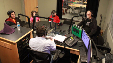 Three youth and an adult wearing headphones sitting behind microphones looking at an adult wearing headphones sitting behind a microphone and a mixing board writing on a piece of paper in a radio studio