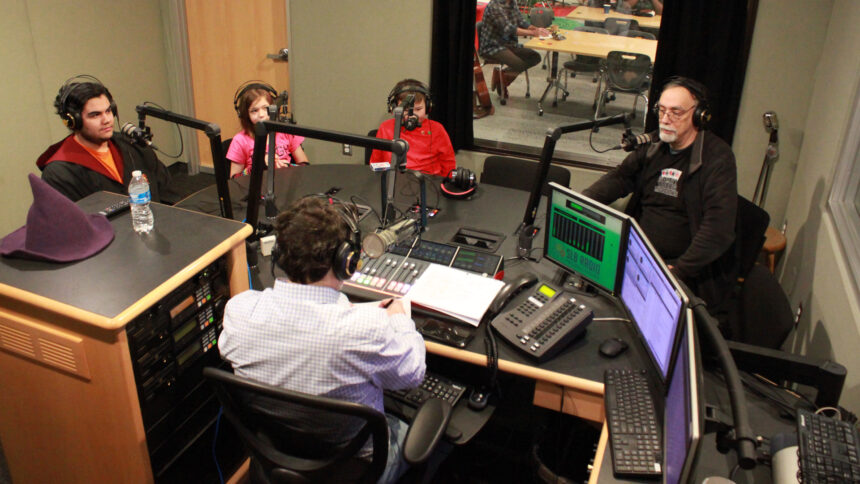 Three youth and an adult wearing headphones sitting behind microphones looking at an adult wearing headphones sitting behind a microphone and a mixing board writing on a piece of paper in a radio studio