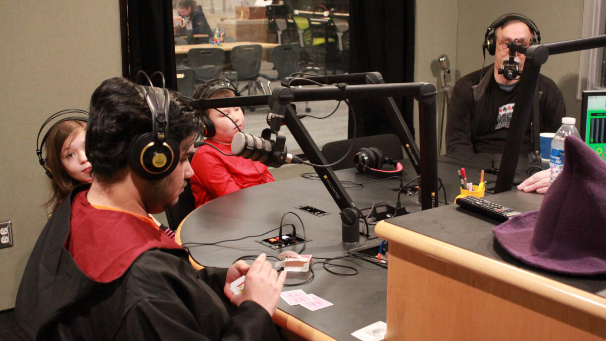 Society-of-American-Magicians-Youth-2 Three youth and an adult wearing headphones sitting behind microphones in a radio studio, one youth interacting with playing cards