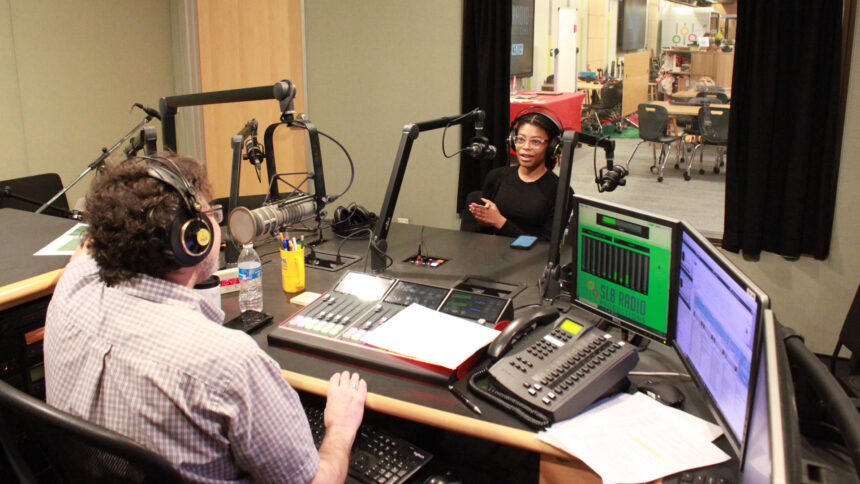 A teen wearing headphones speaking into a microphone being interviewed by an adult wearing headphones sitting behind a microphone and a mixing board in a radio studio