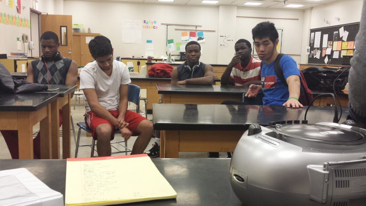 Five youth looking at a cd player and sitting at classroom desks