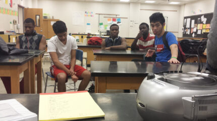 Five youth looking at a cd player and sitting at classroom desks