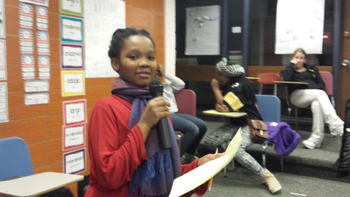 A youth holding a piece of paper and speaking into a microphone in a classroom