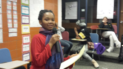 A youth holding a piece of paper and speaking into a microphone in a classroom