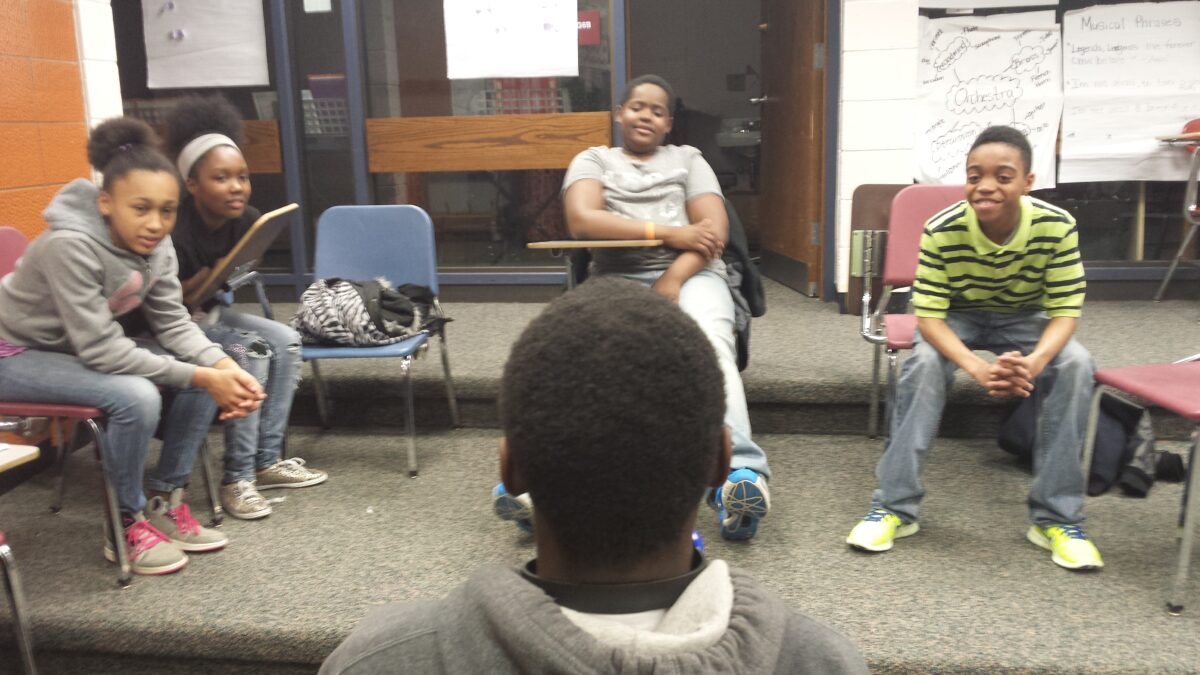 A youth facing four youth sitting in classroom chairs