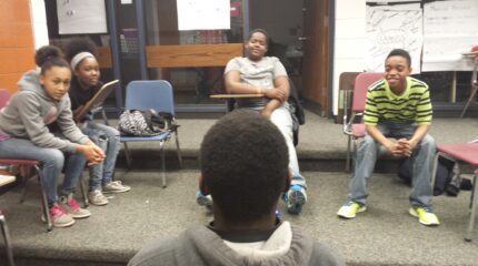 A youth facing four youth sitting in classroom chairs