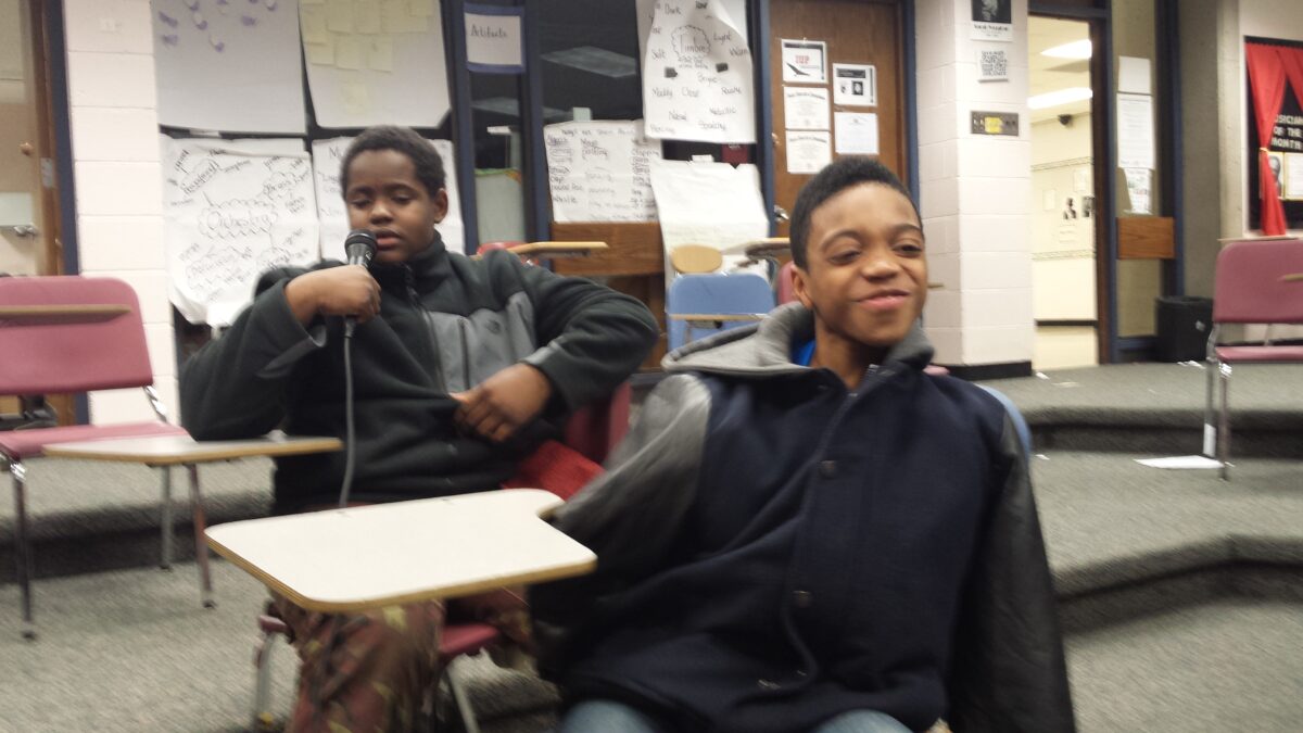 Two youth sitting at classroom desks, one holding and speaking into a microphone