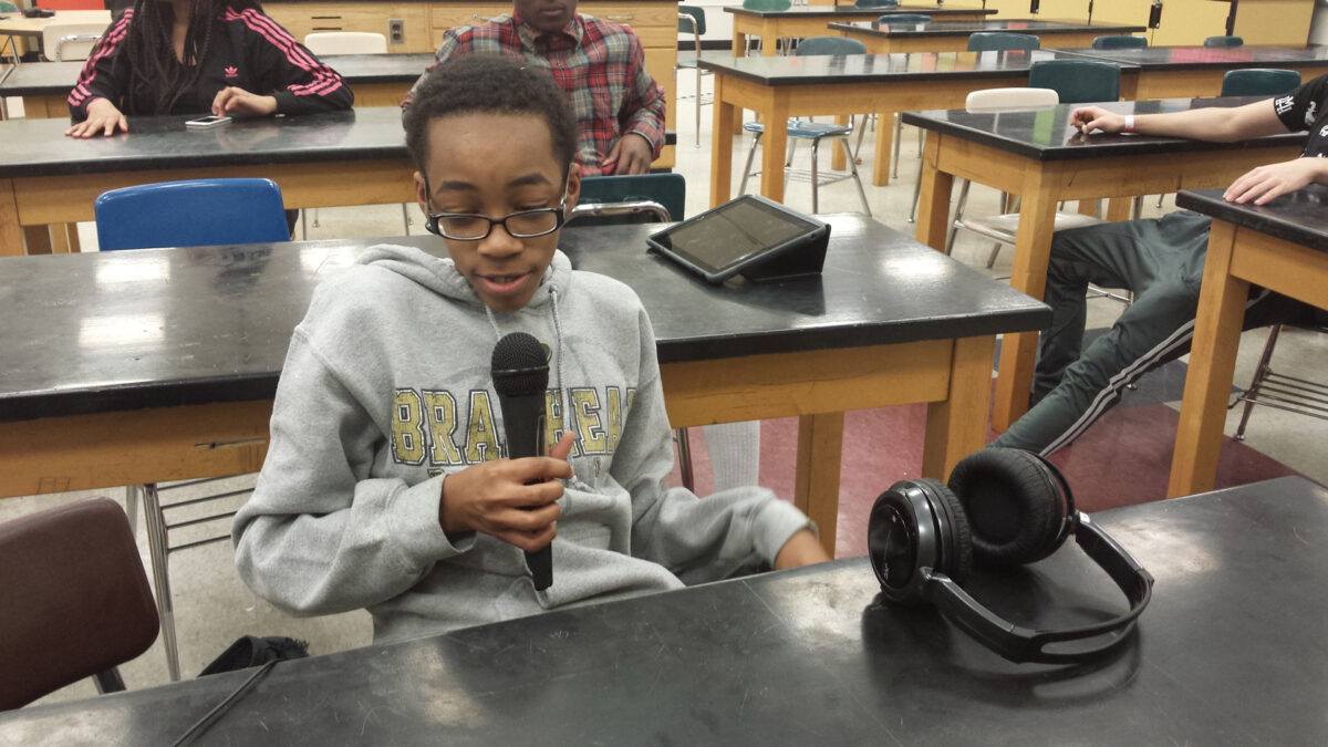 A teen holding and speaking into a microphone sitting at a classroom desk
