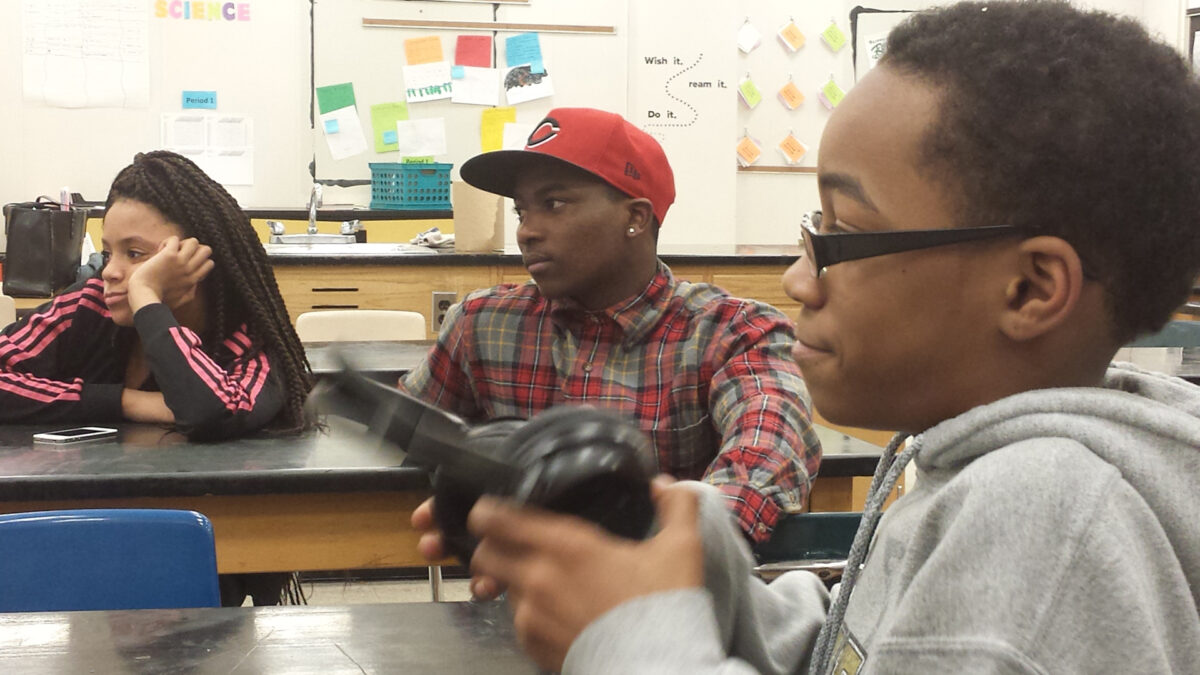 Three teens looking to the left sitting at classroom desks, one teen holding headphones