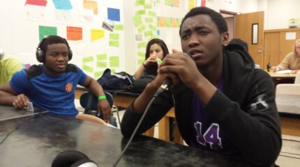 Three teens sitting at classroom desks, one holding and speaking into a microphone