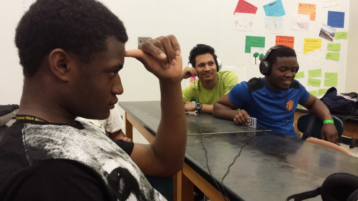 Four teens sitting around a classroom desk, two wearing headphones