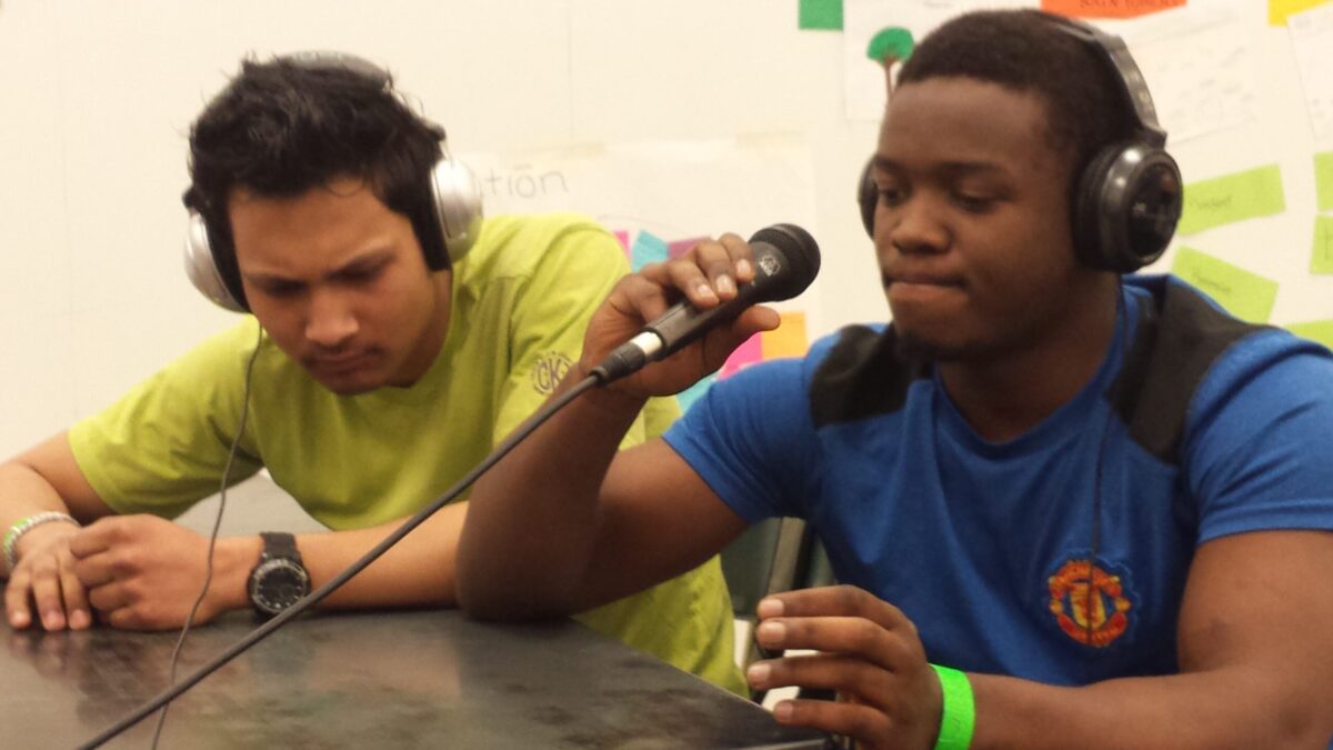 Two teens wearing headphones sitting at a classroom desk, one holding a microphone