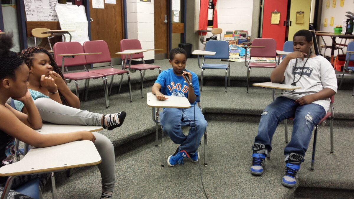 Four youth sitting at classroom desks, one holding and speaking into a microphone