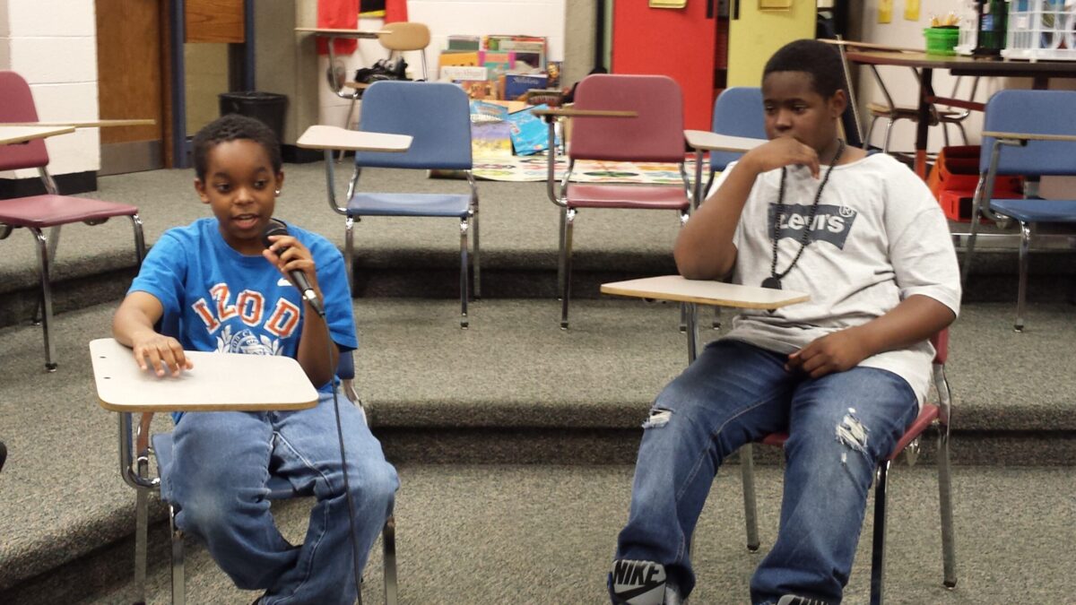 Two youth sitting at classroom desks, one holding and speaking into a microphone