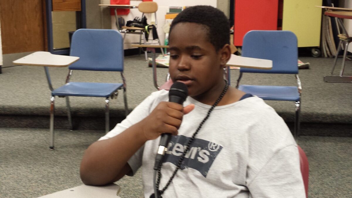A youth holding and speaking into a microphone sitting at a classroom desk