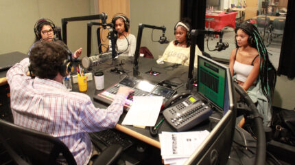 Three teens and an adult wearing headphones speaking into microphones being interviewed by an adult wearing headphones sitting behind a microphone and a mixer in a radio studio