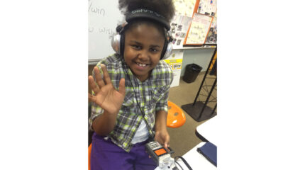 A youth wearing headphone holding a portable recorder and waving to the camera while sitting in front of a whiteboard in a classroom