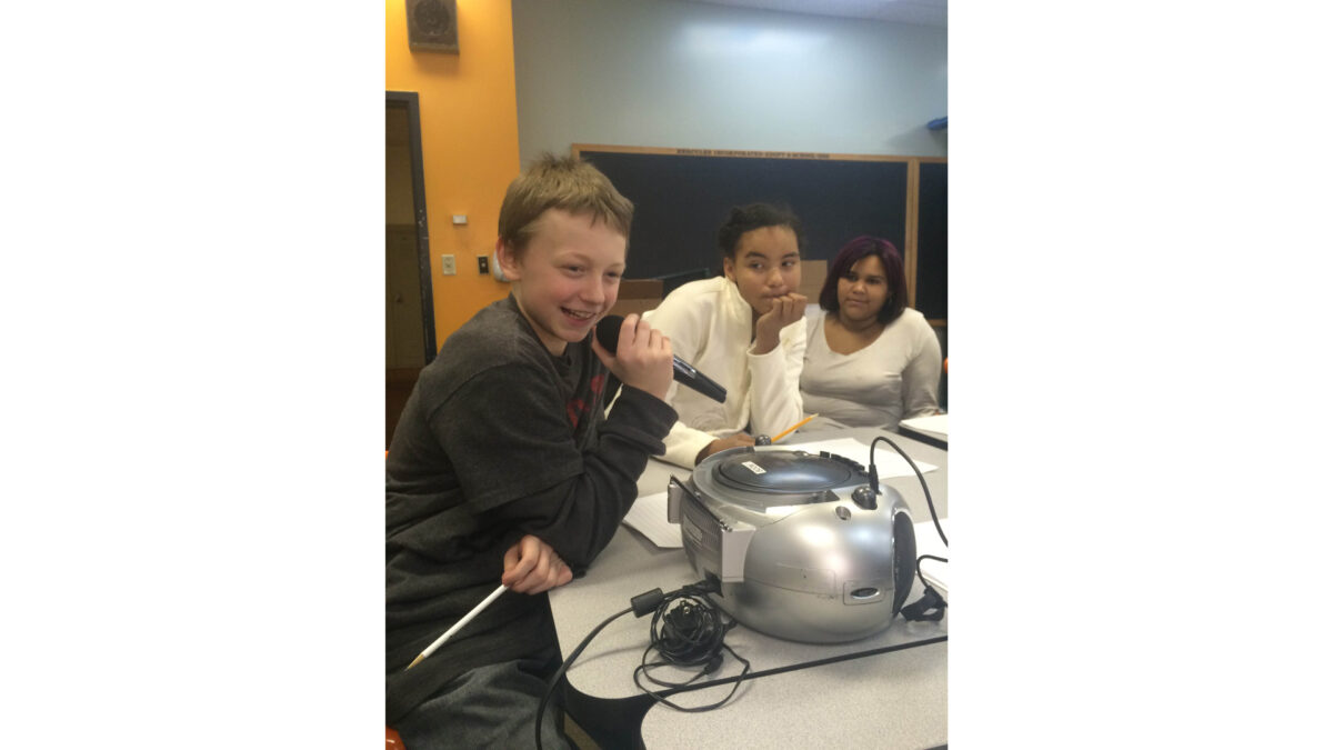 Two youth looking at another youth holding and speaking into a microphone, all sitting behind a cd player on a gray desk in a classroom