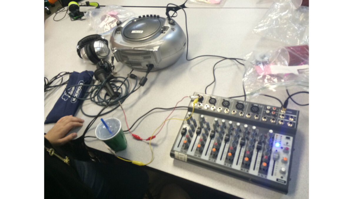 A cd player, two pair of headphones, a microphone, a green plastic cup with a straw, and a mixer, all sitting on a light gray table
