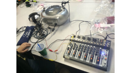 A cd player, two pair of headphones, a microphone, a green plastic cup with a straw, and a mixer, all sitting on a light gray table
