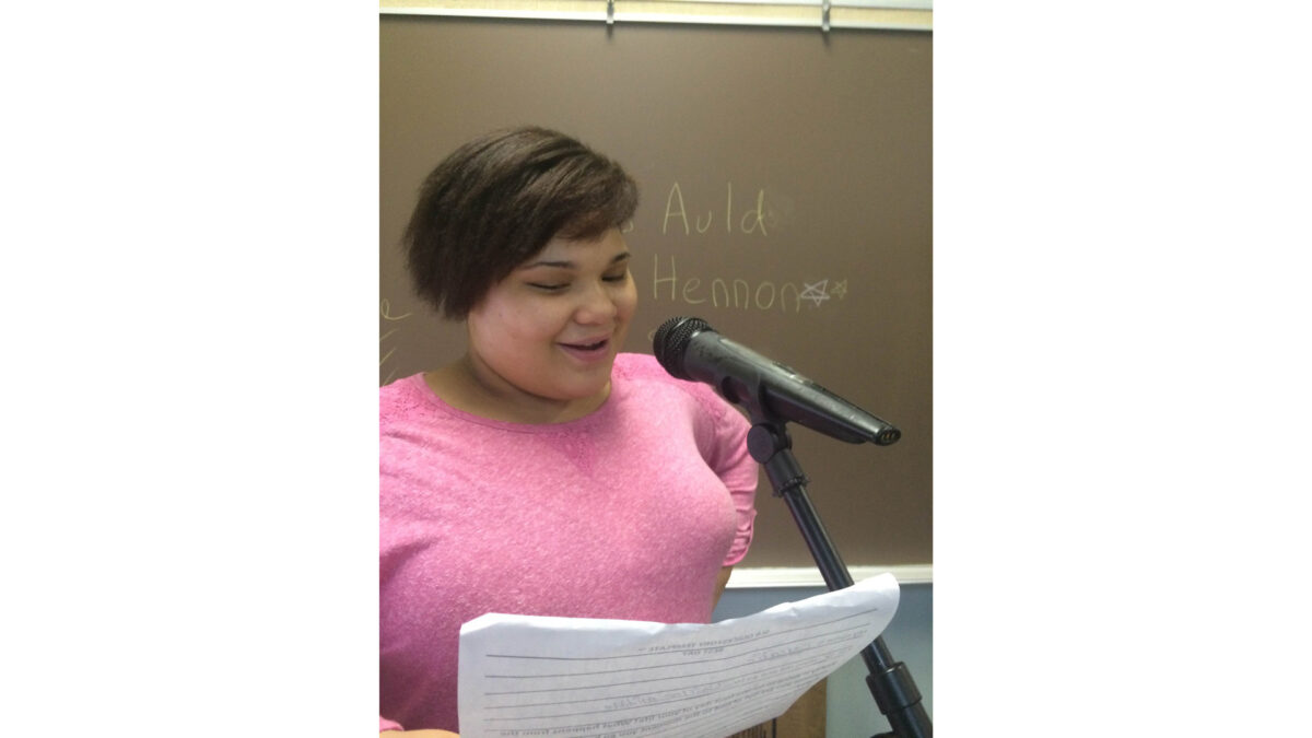 A youth holding a piece of paper and speaking into a microphone standing in front of a blackboard in a classroom
