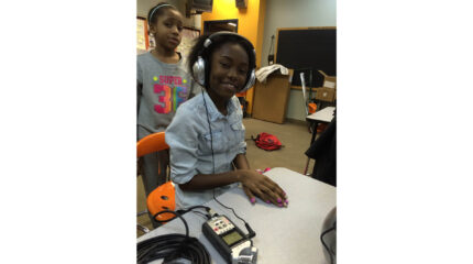 A youth standing behind another youth sitting while wearing headphones connected to a portable recorder on a desk in a classroom