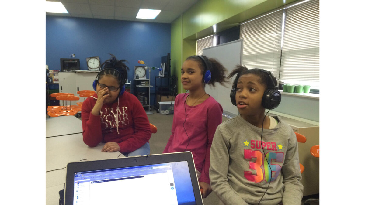 Three youth wearing headphones looking to the right sitting behind a laptop computer on a white desk in a classroom