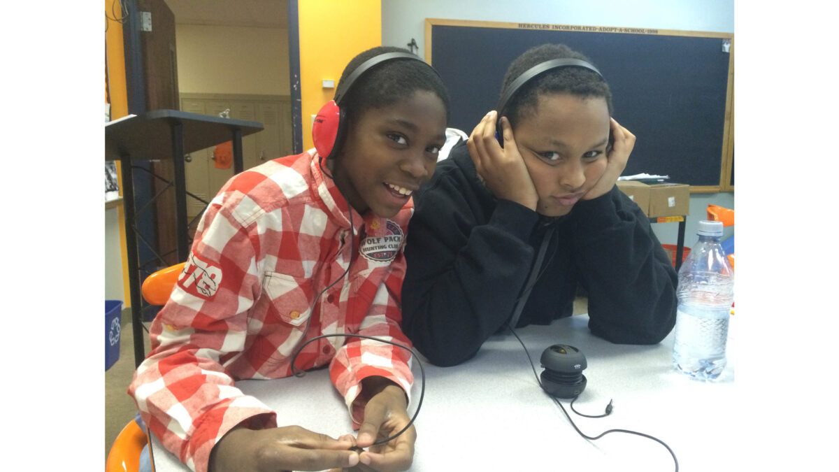 Two youth wearing headphones smiling at the camera while sitting at a white desk in a classroom